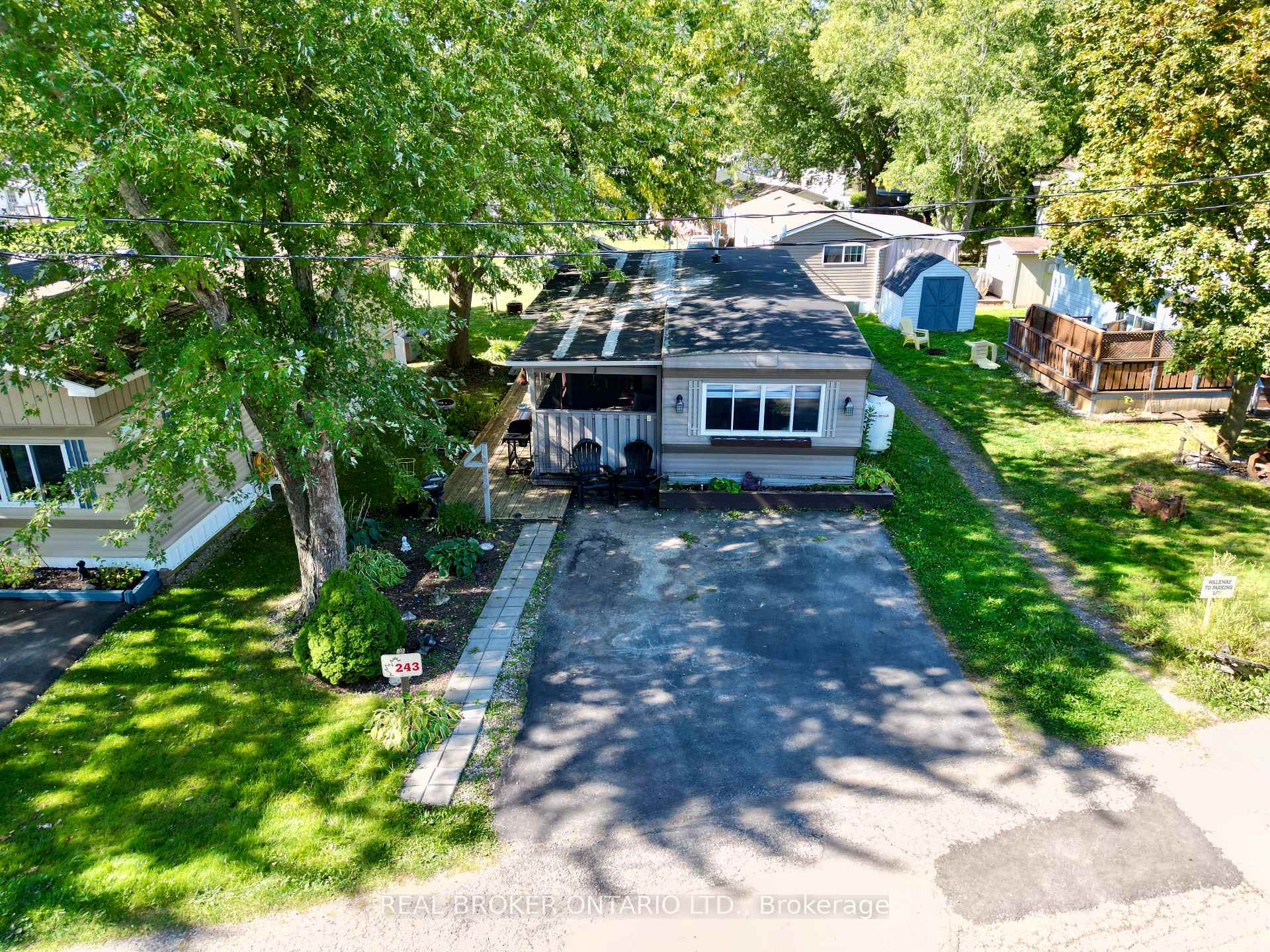 Aerial view of home 243 with a wide driveway, front deck, mature trees, and a blue shed.