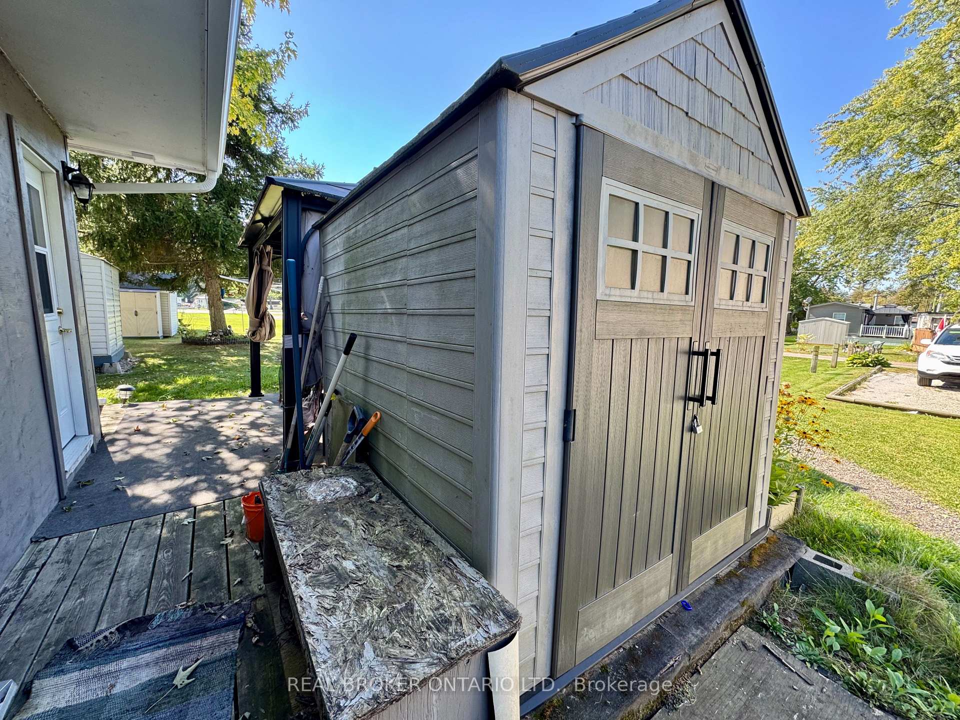 Large gray garden shed with double doors and small windows beside a deck and sunny yard.