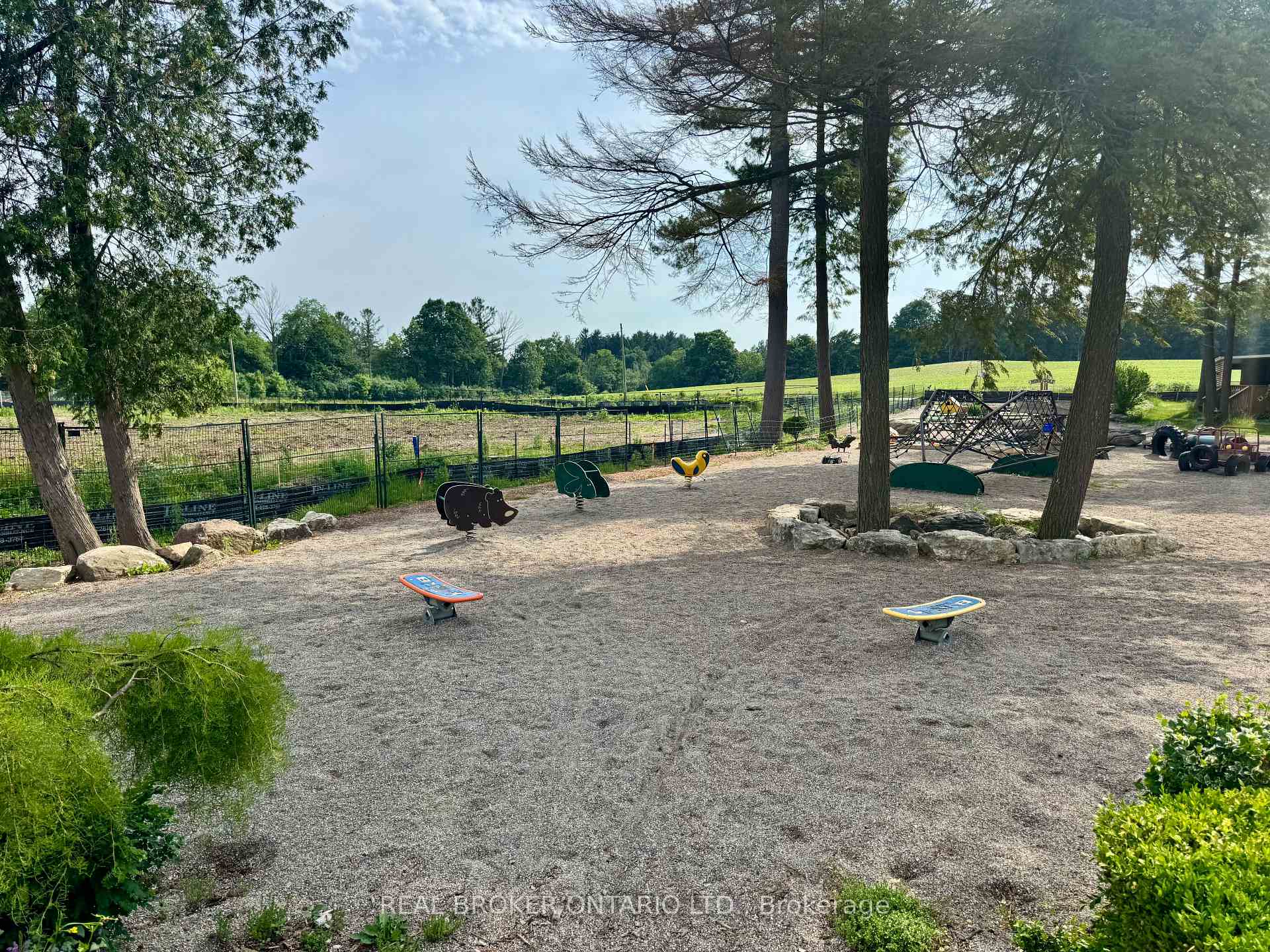 Gravel playground with small play structures and climbing equipment beneath mature trees.