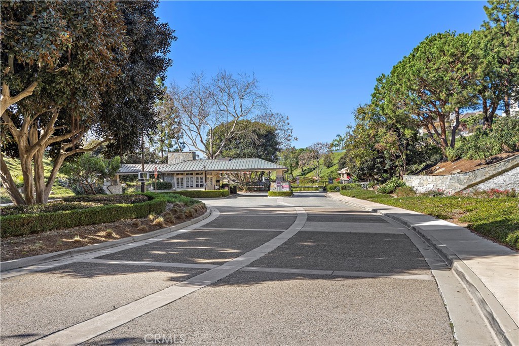 Gated community entrance with a low guardhouse and curved roadway beneath mature trees.