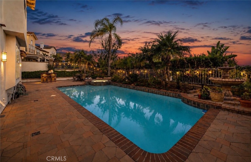 Backyard pool and paved patio with palm trees and garden lights at dusk.