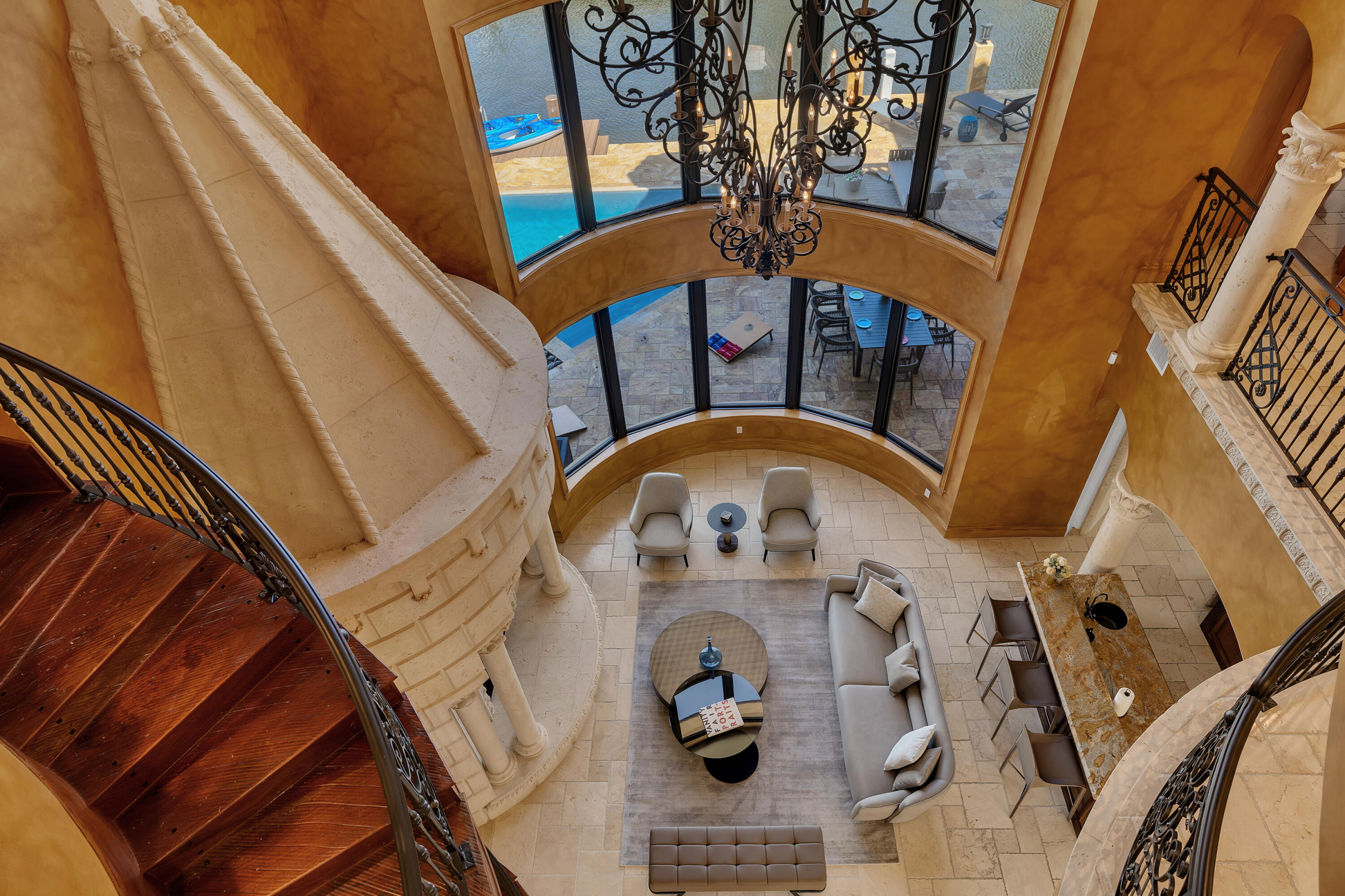 Grand living room seen from above, with a curved staircase, chandelier, and pool-view windows.
