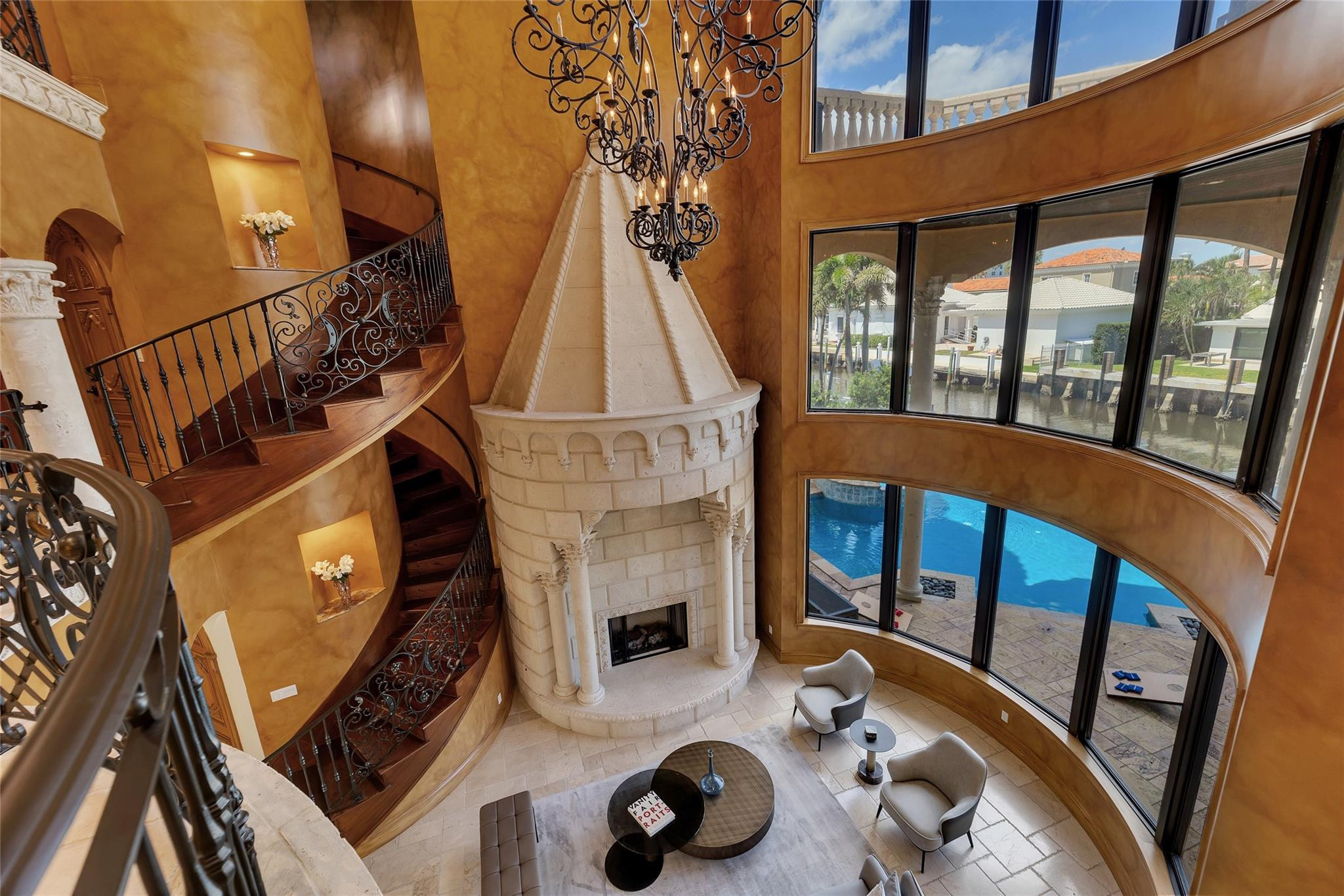 Two-story living room with a sculpted stone fireplace, curved staircase, and windows overlooking a pool and canal.