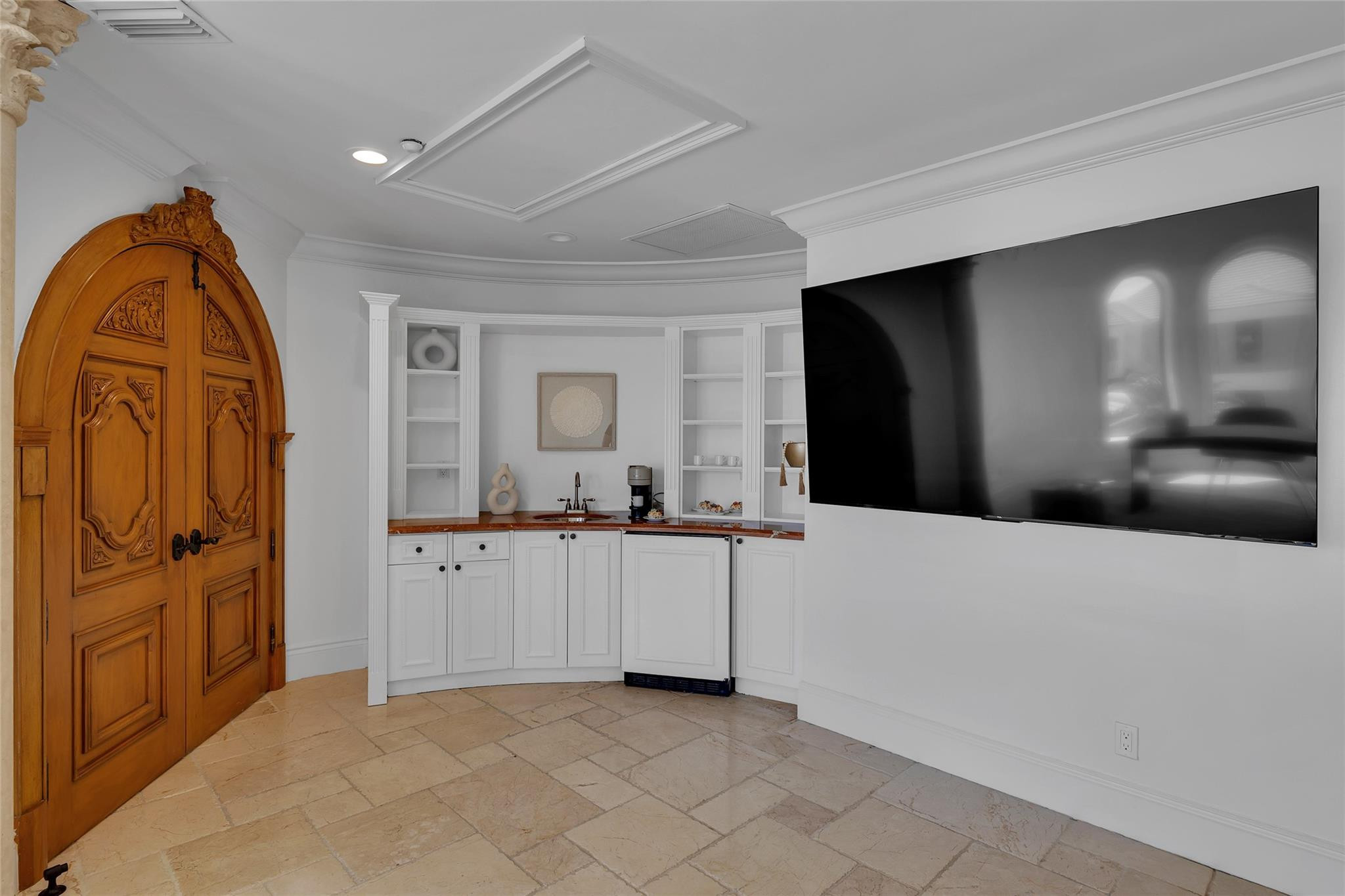 Room with an ornate arched wooden door, built-in kitchenette, stone tile floor, and large wall-mounted television.