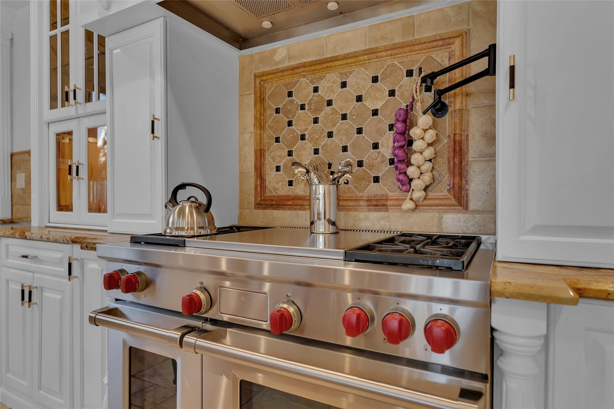 Kitchen range with red knobs, tan stone backsplash, and hanging garlic beside white cabinetry.