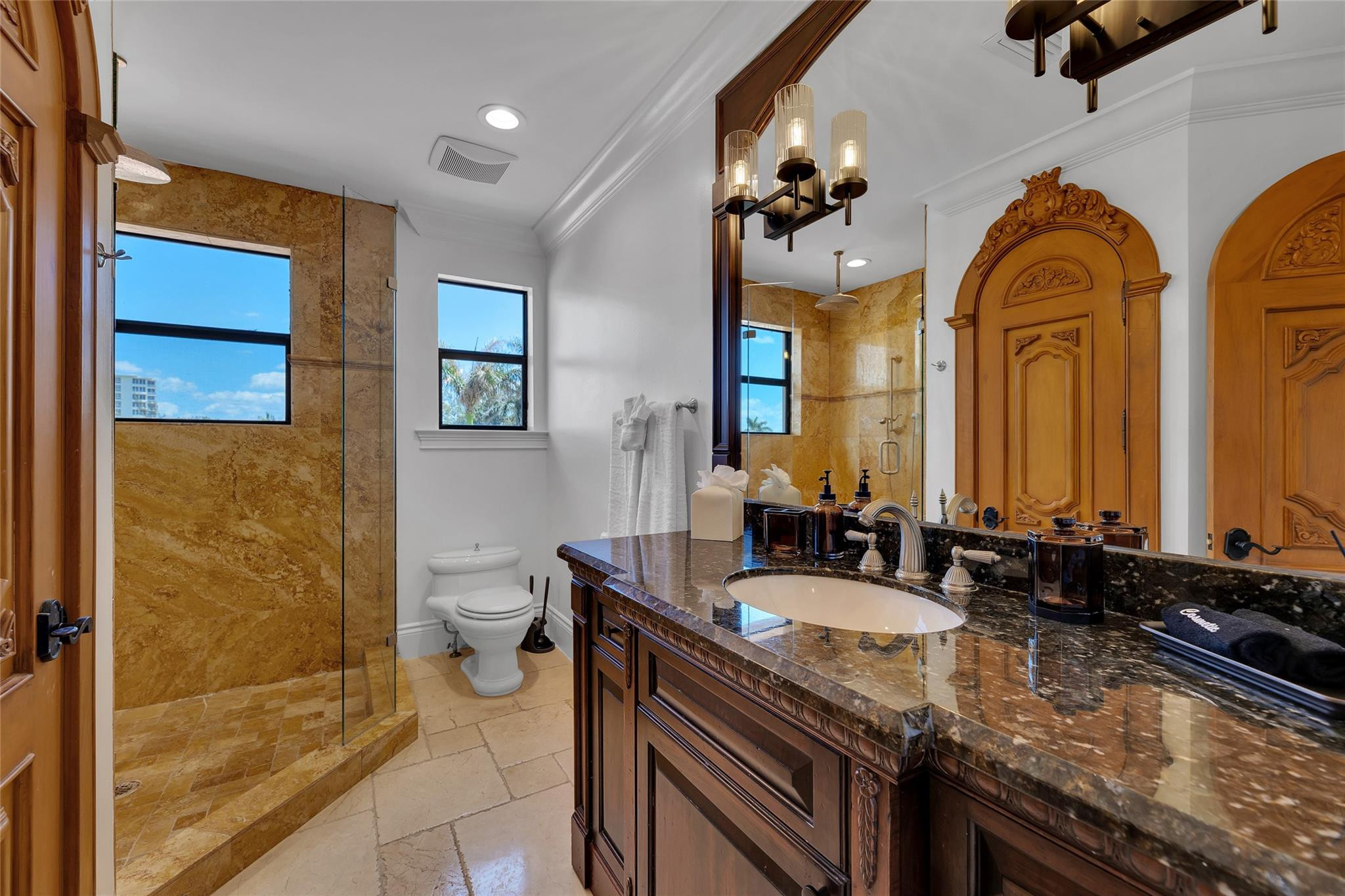 Bathroom with a golden stone shower, ornate wood vanity, and two daylight-filled windows.