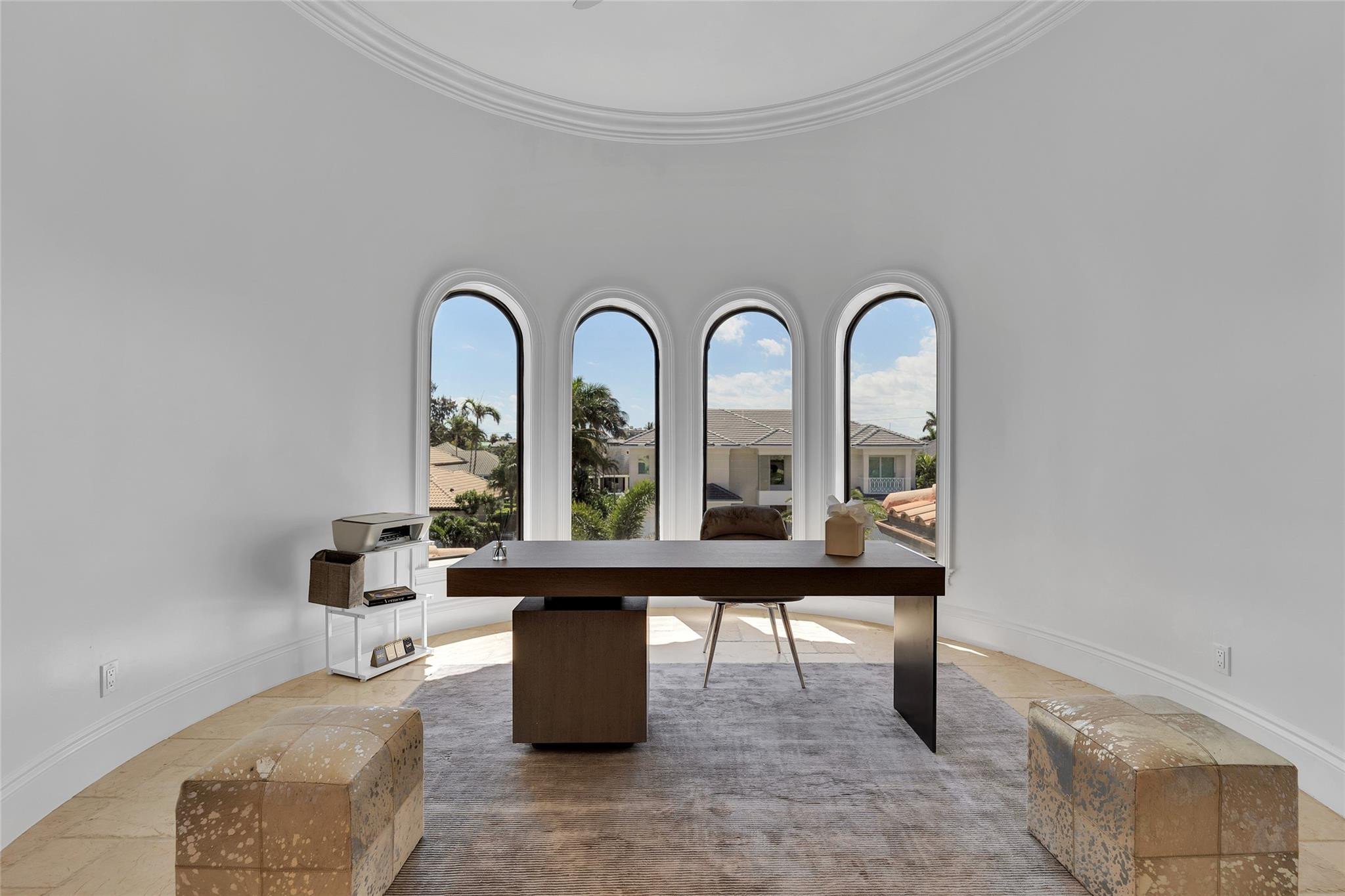 Furnished home office with a dark wood desk and four arched windows overlooking palm trees.