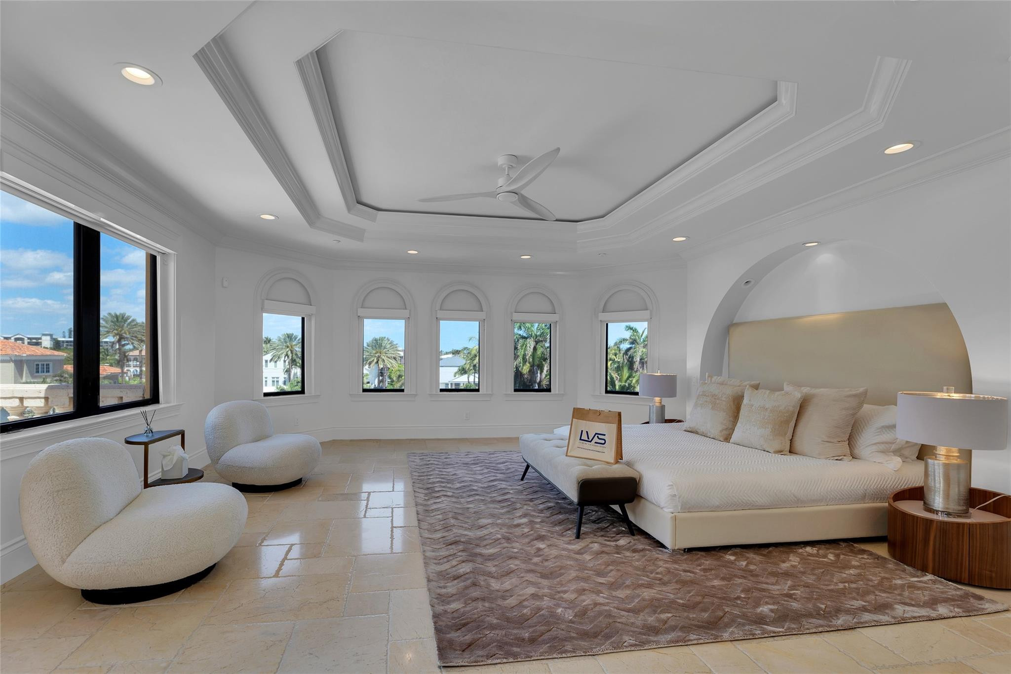 Staged bedroom with a large bed, arched windows, and two rounded lounge chairs.