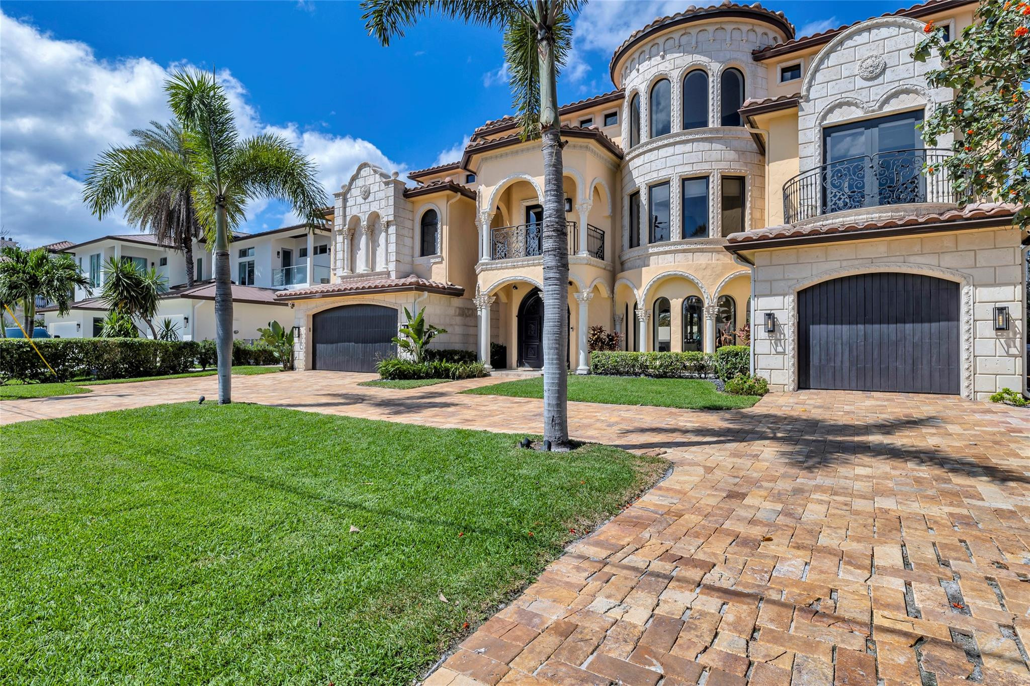 Mediterranean-style stone and stucco home with arched balconies, palm trees, and a broad paver driveway.