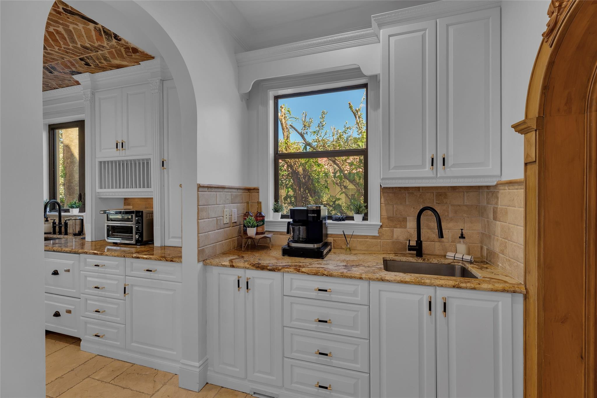 Kitchen with white cabinetry, tan stone counters, and a black sink beneath a window overlooking trees.