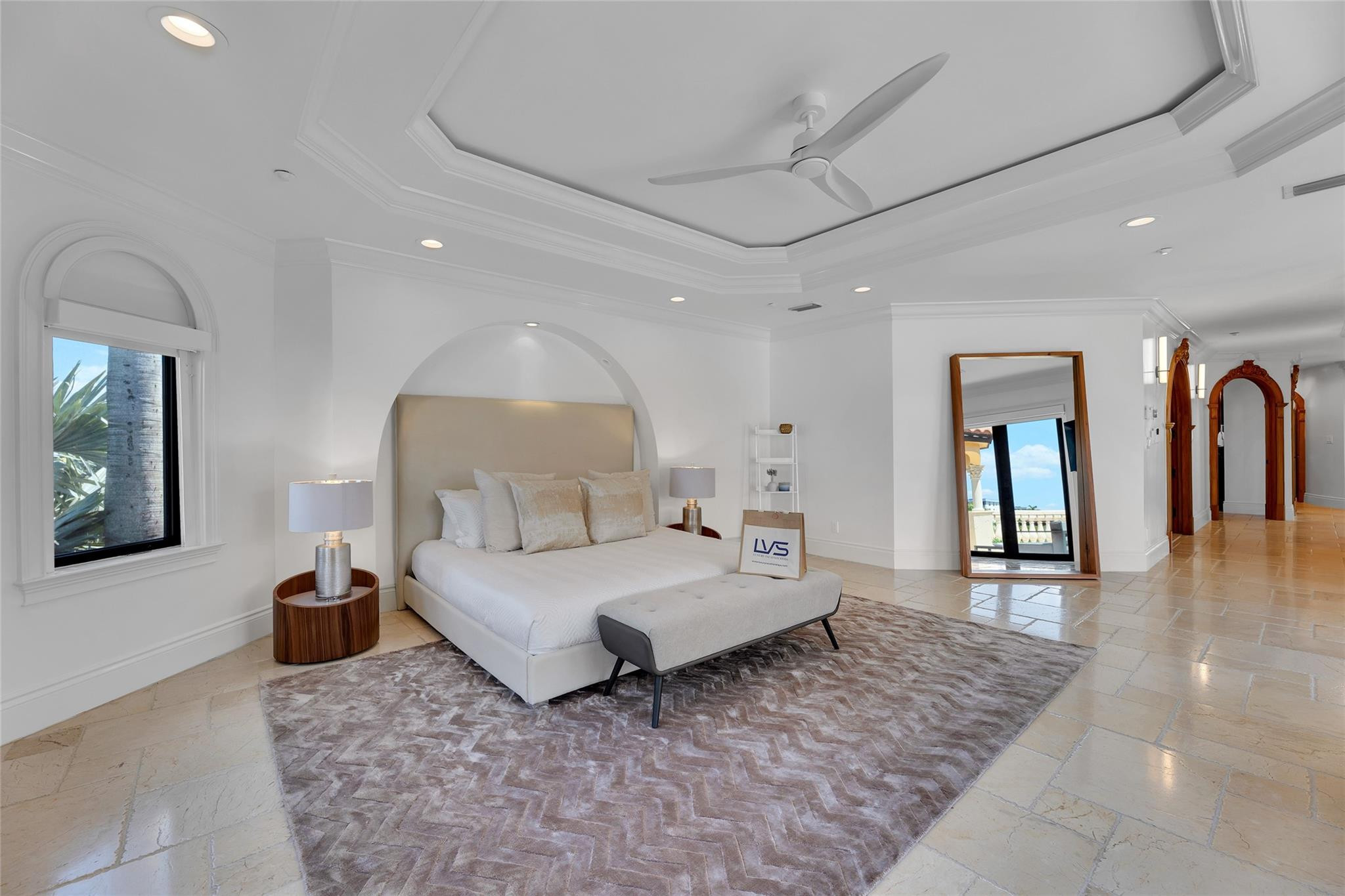 Bedroom with a white bed in an arched niche, patterned rug, and tall mirror reflecting a balcony doorway.
