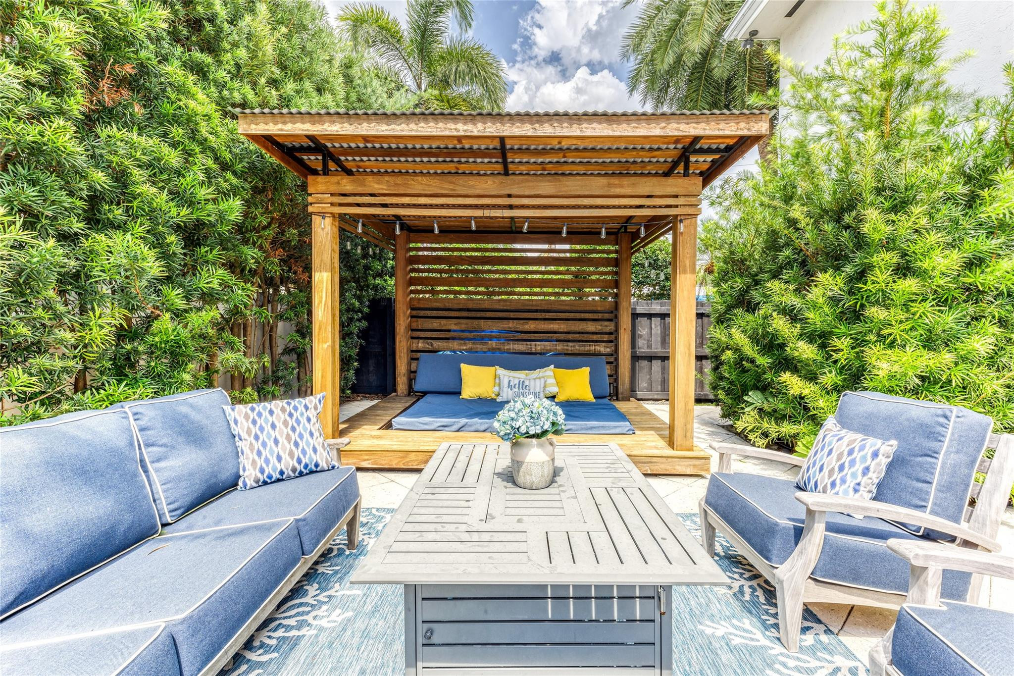 Outdoor patio with a wooden pergola daybed, blue seating, and lush tropical greenery.