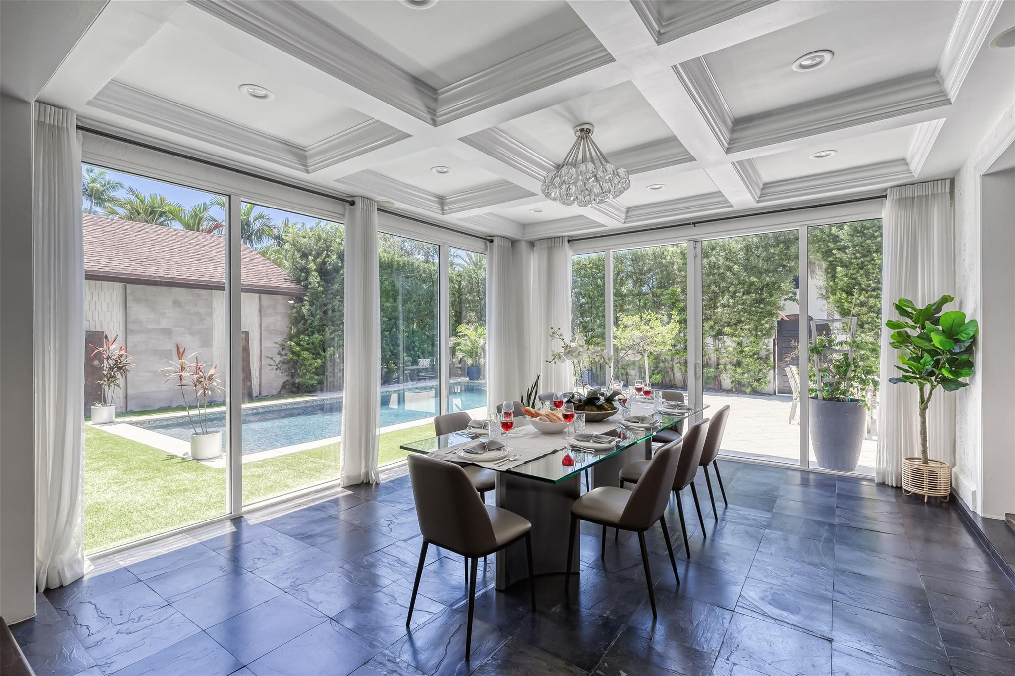 Dining room with a glass table, coffered ceiling, and floor-to-ceiling windows overlooking the pool.