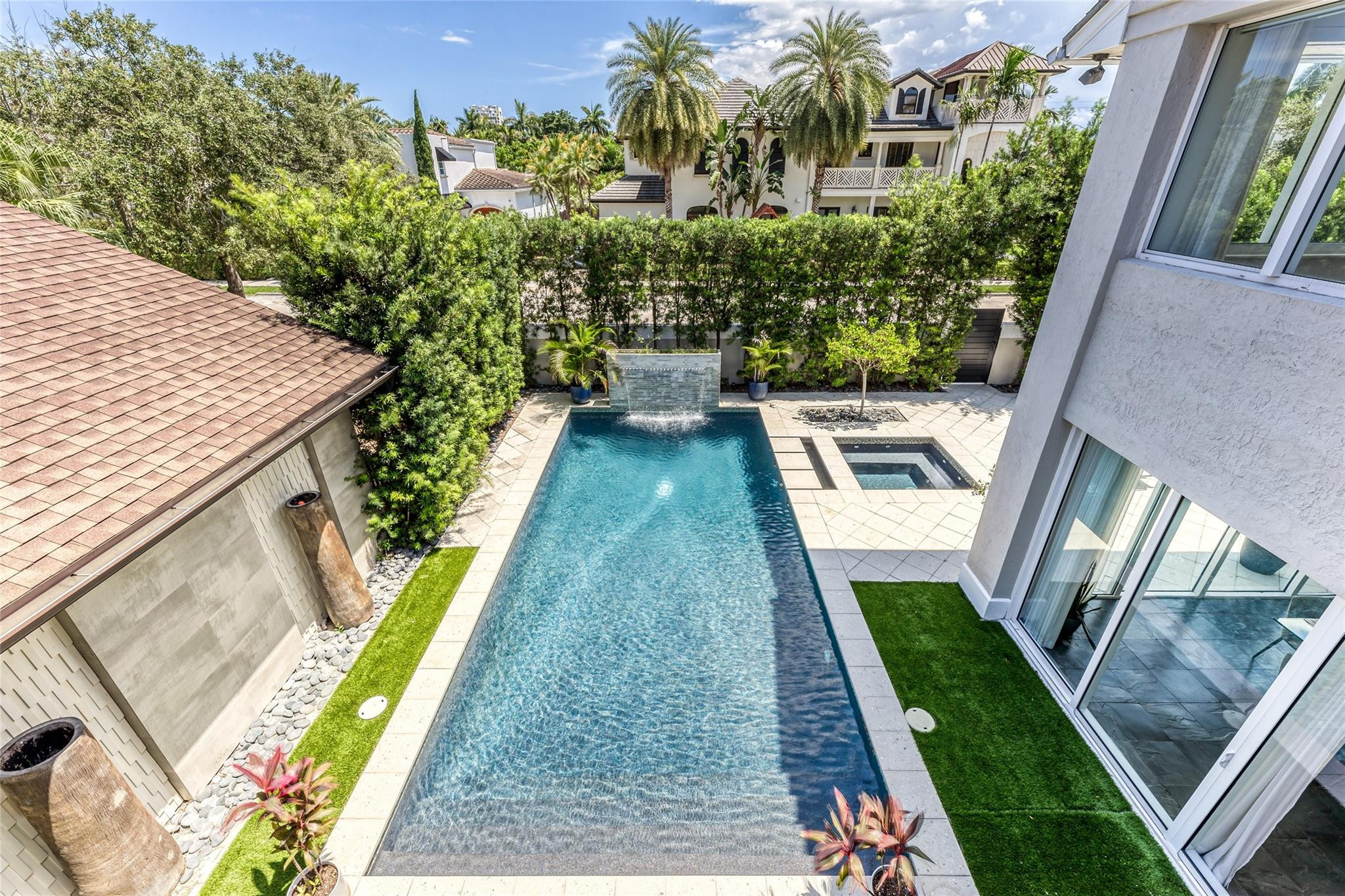 Rectangular courtyard pool with a raised waterfall feature, spa, stone deck, and tropical landscaping.