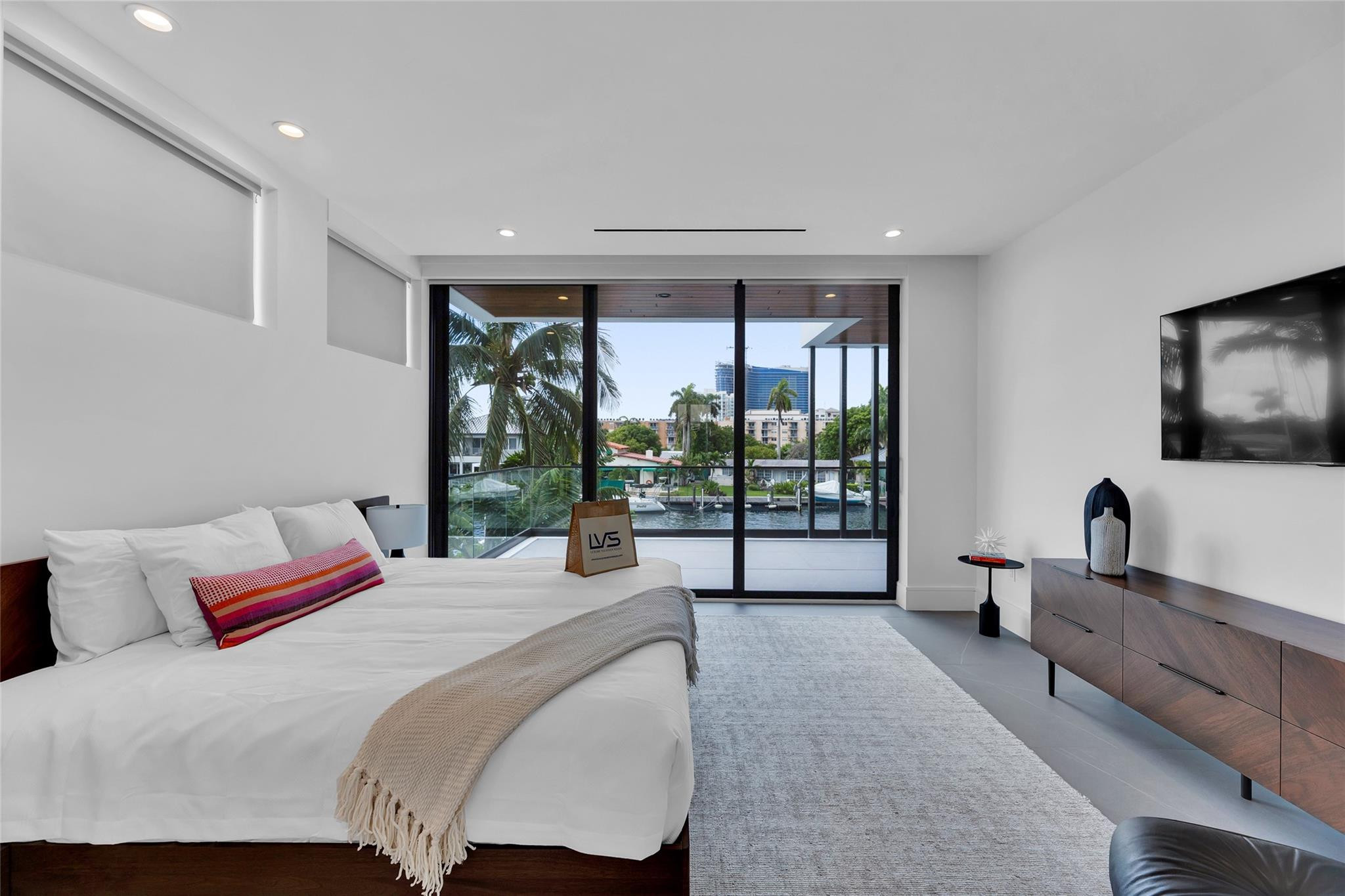 Modern bedroom with a white bed and sliding glass doors overlooking a palm-lined canal.