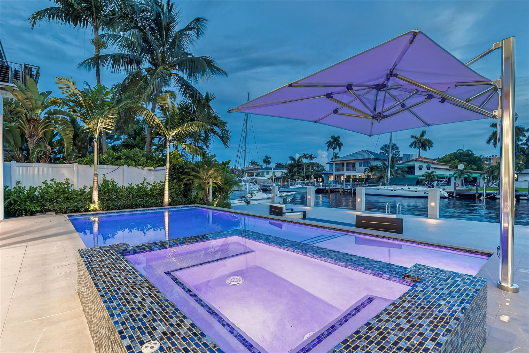Waterfront pool and tiled spa under a purple umbrella, with docked boats and palm trees along the canal.