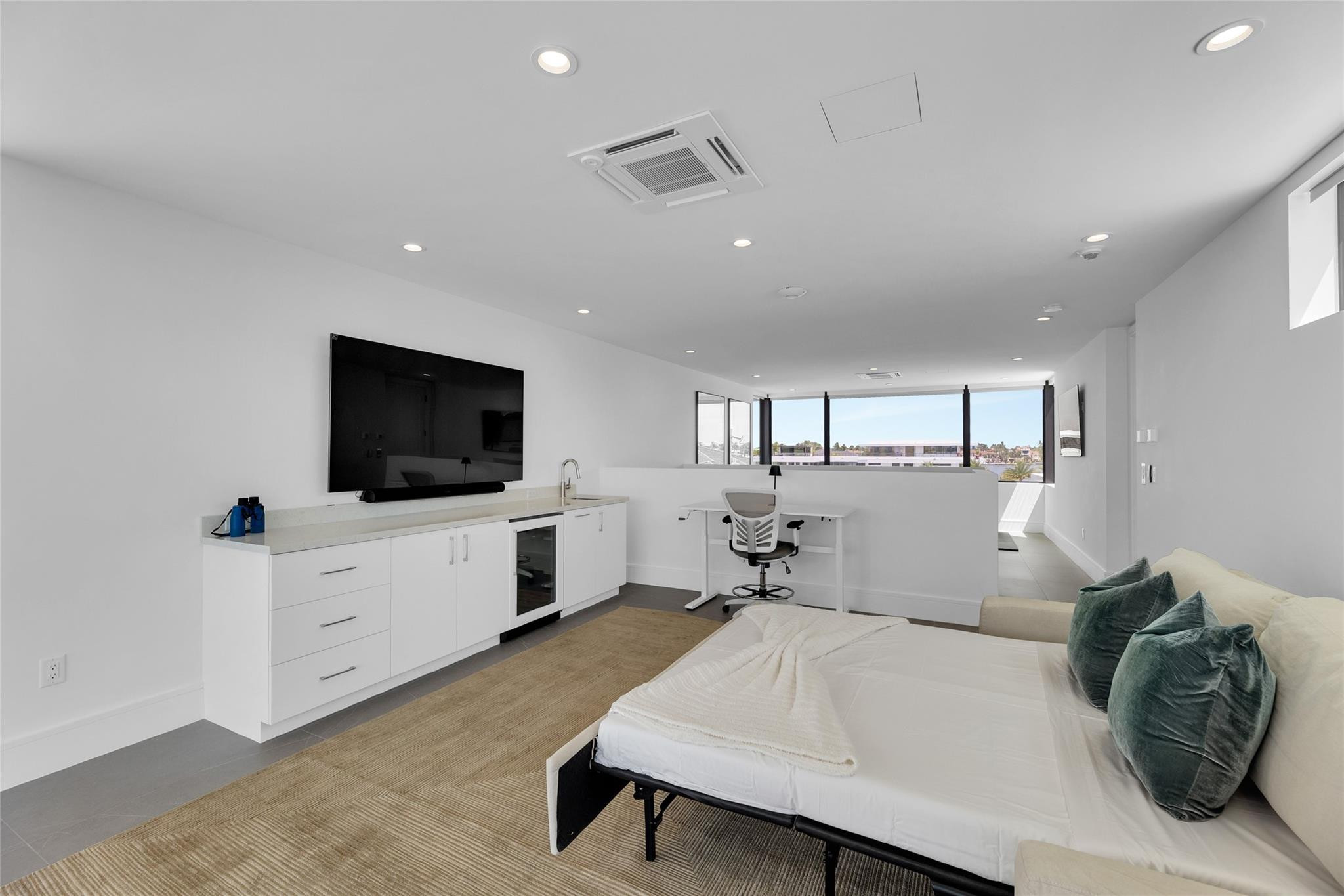 Modern bedroom with a white bed, teal cushions, built-in cabinets, and a desk by broad windows.