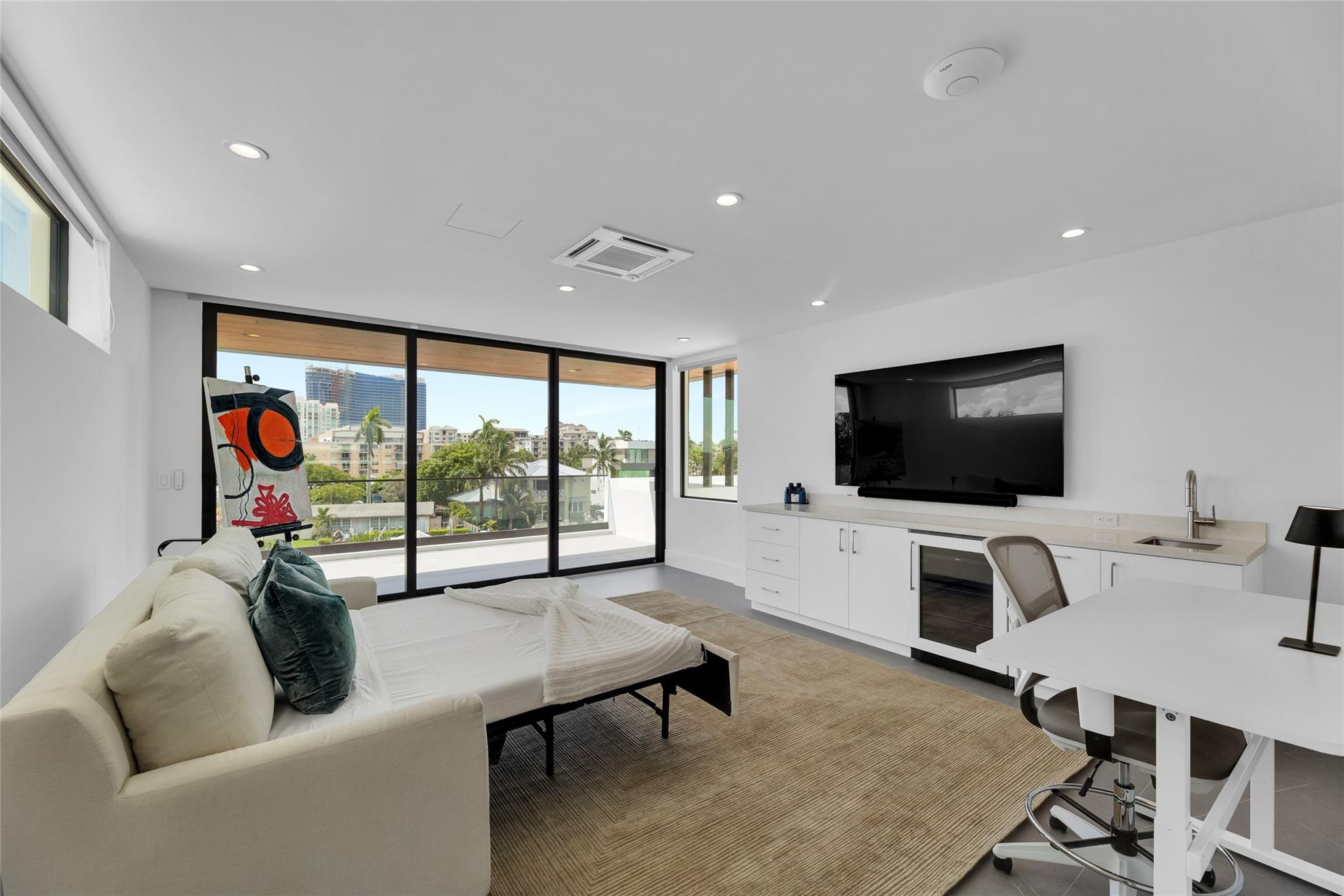 Modern studio with a sofa-bed, built-in desk, and large windows overlooking city buildings.