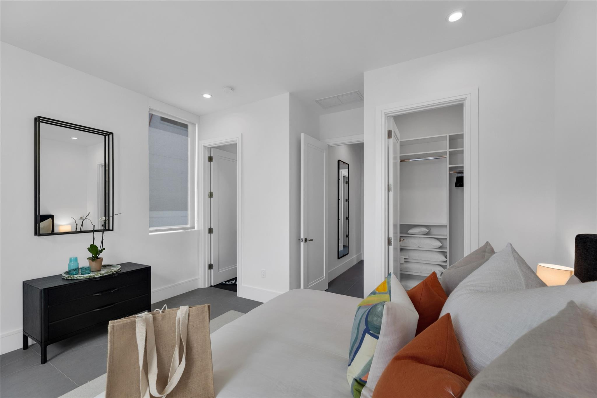 Modern bedroom with white bedding, colorful accent pillows, black dresser, and open built-in closet.