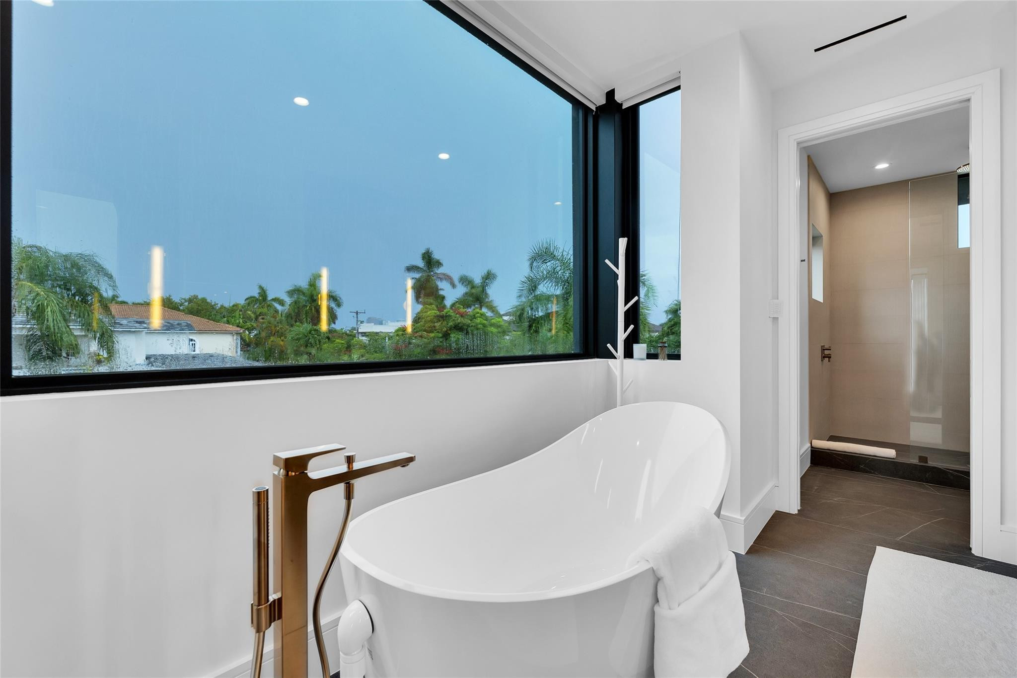 Modern bathroom with a freestanding tub beside a large window overlooking palm trees and an adjoining tiled shower.