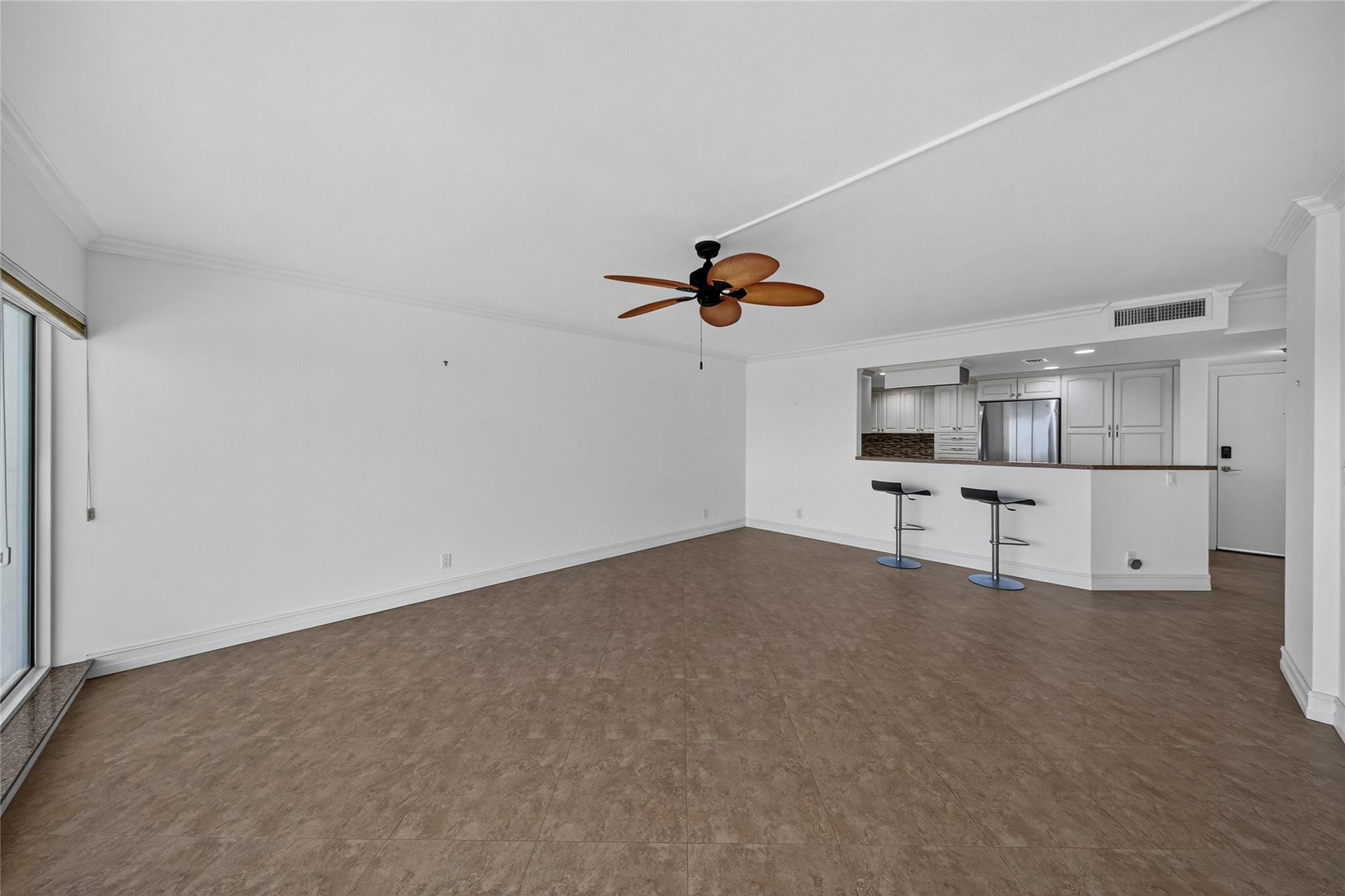 Vacant living area with brown tile floors, ceiling fan, and kitchen pass-through with bar stools.