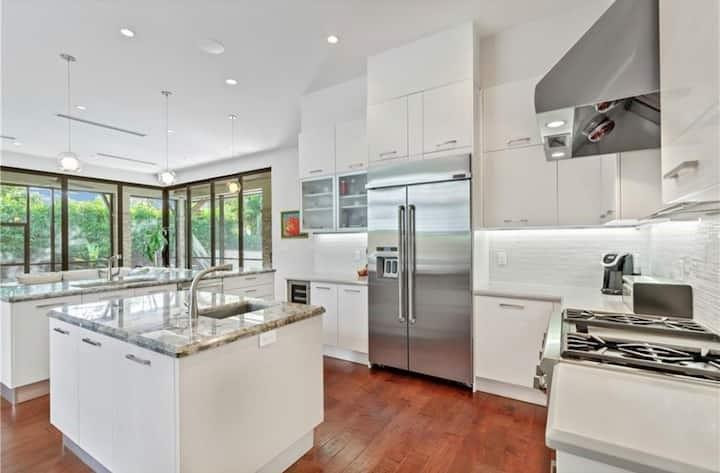 Bright white kitchen with a stone island, stainless steel appliances, hardwood floors, and windows overlooking greenery.