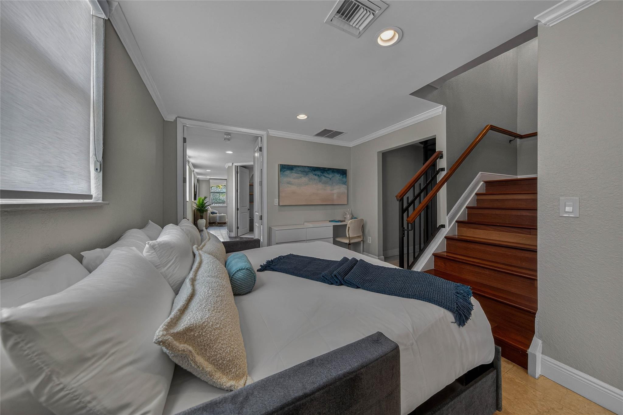 Furnished bedroom with a white bed beside a wooden staircase with black railings.