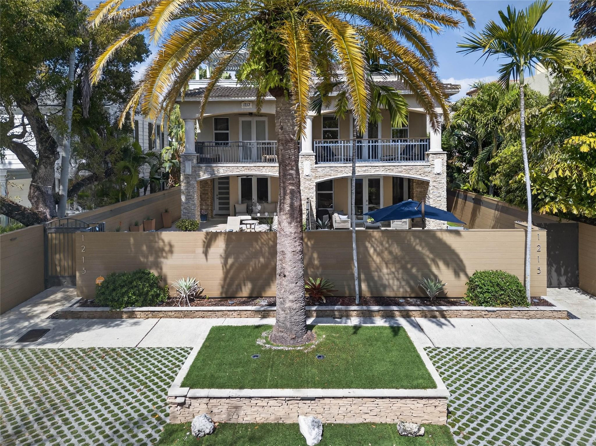 Two-story home with a stone-accented facade, deep balcony, tropical landscaping, and a fenced patio.