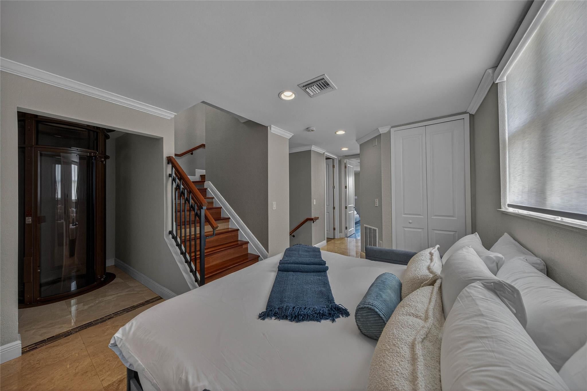 Staged bedroom with a white bed, blue throws, wood staircase, and glass elevator.