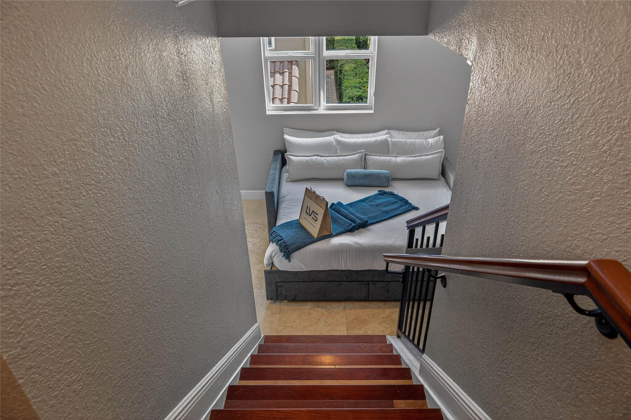 Bedroom at the foot of a staircase, with a white bed, blue throws, and a window.