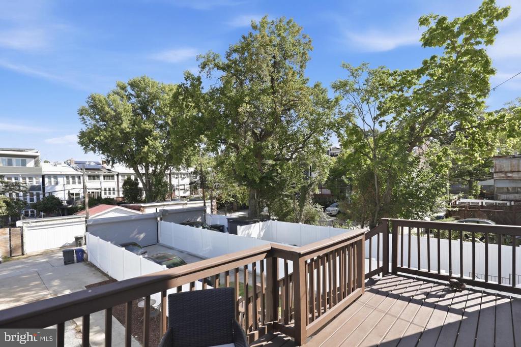 Wooden deck with railings and a chair overlooking a fenced parking area and leafy trees.