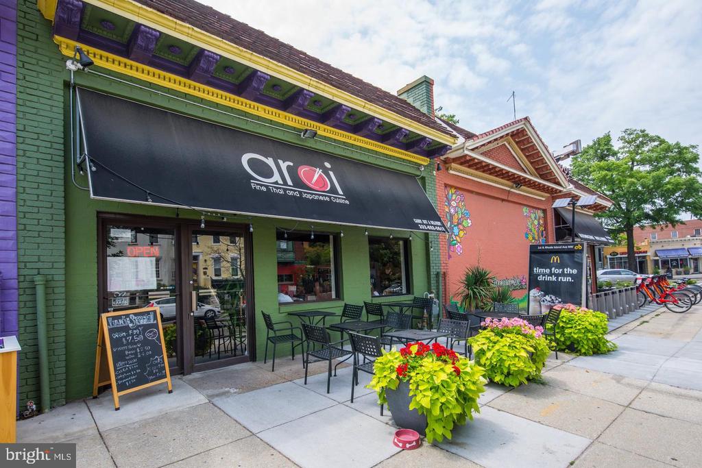 Colorful restaurant storefront with outdoor patio seating, flower planters, and a neighboring mural.