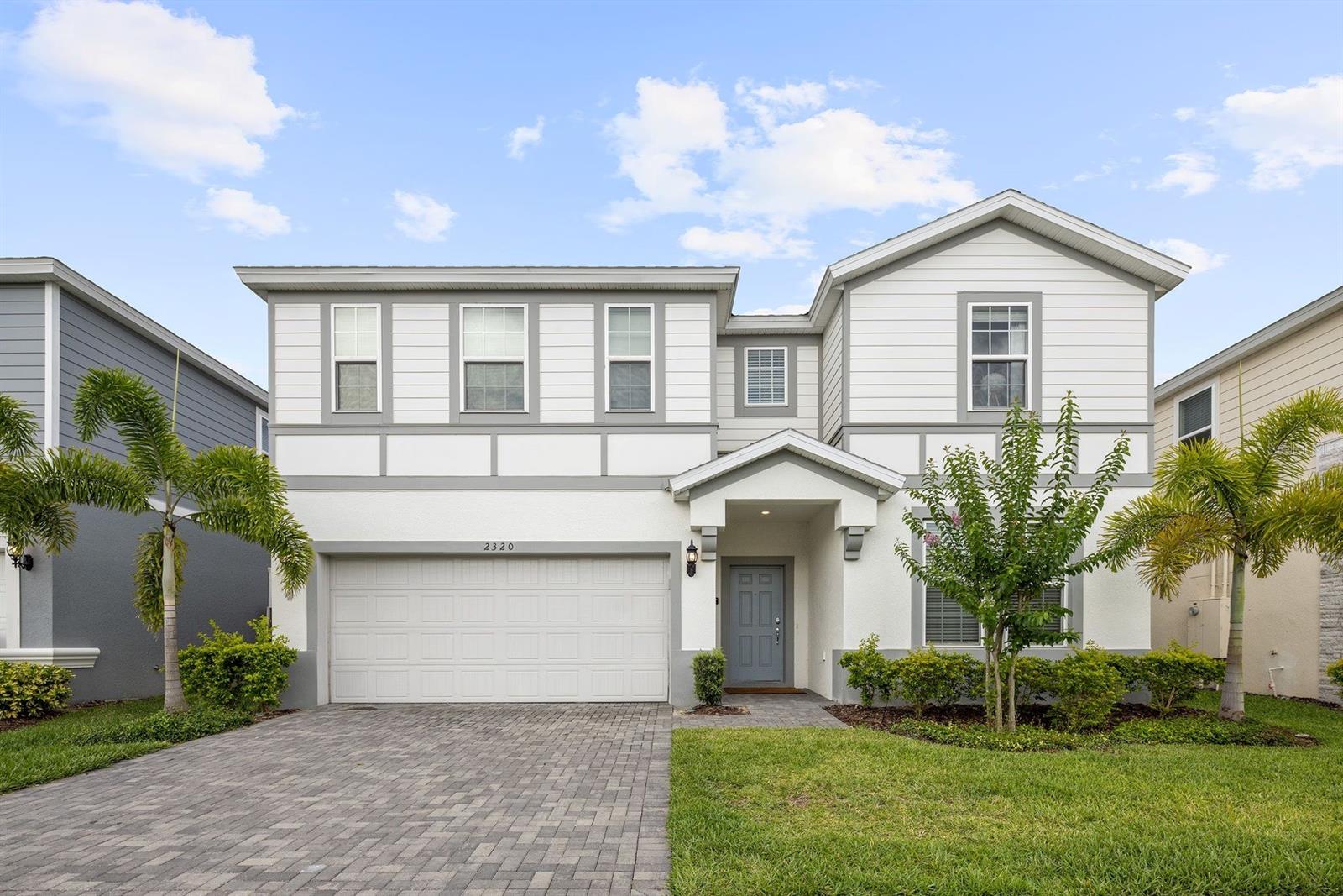 Two-story white house with a double garage, paver driveway, covered entry, and palm trees.