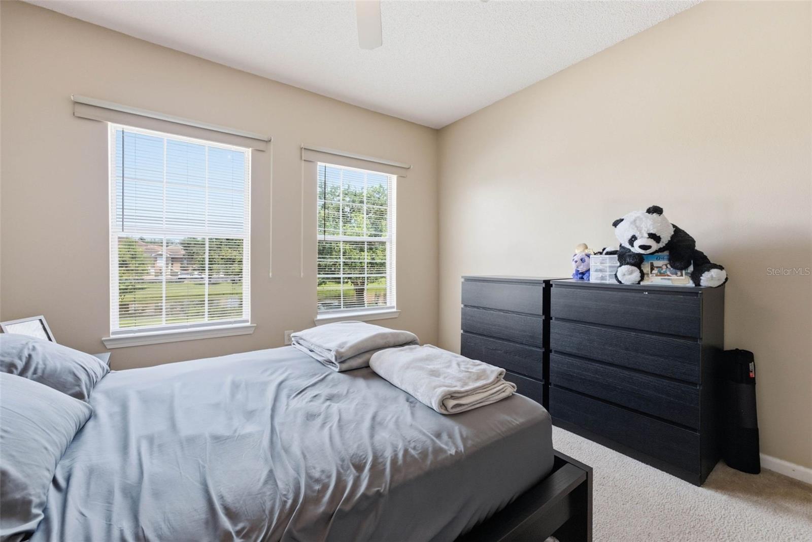 Bedroom with a gray bed, two bright windows, and a dark dresser topped with stuffed animals.