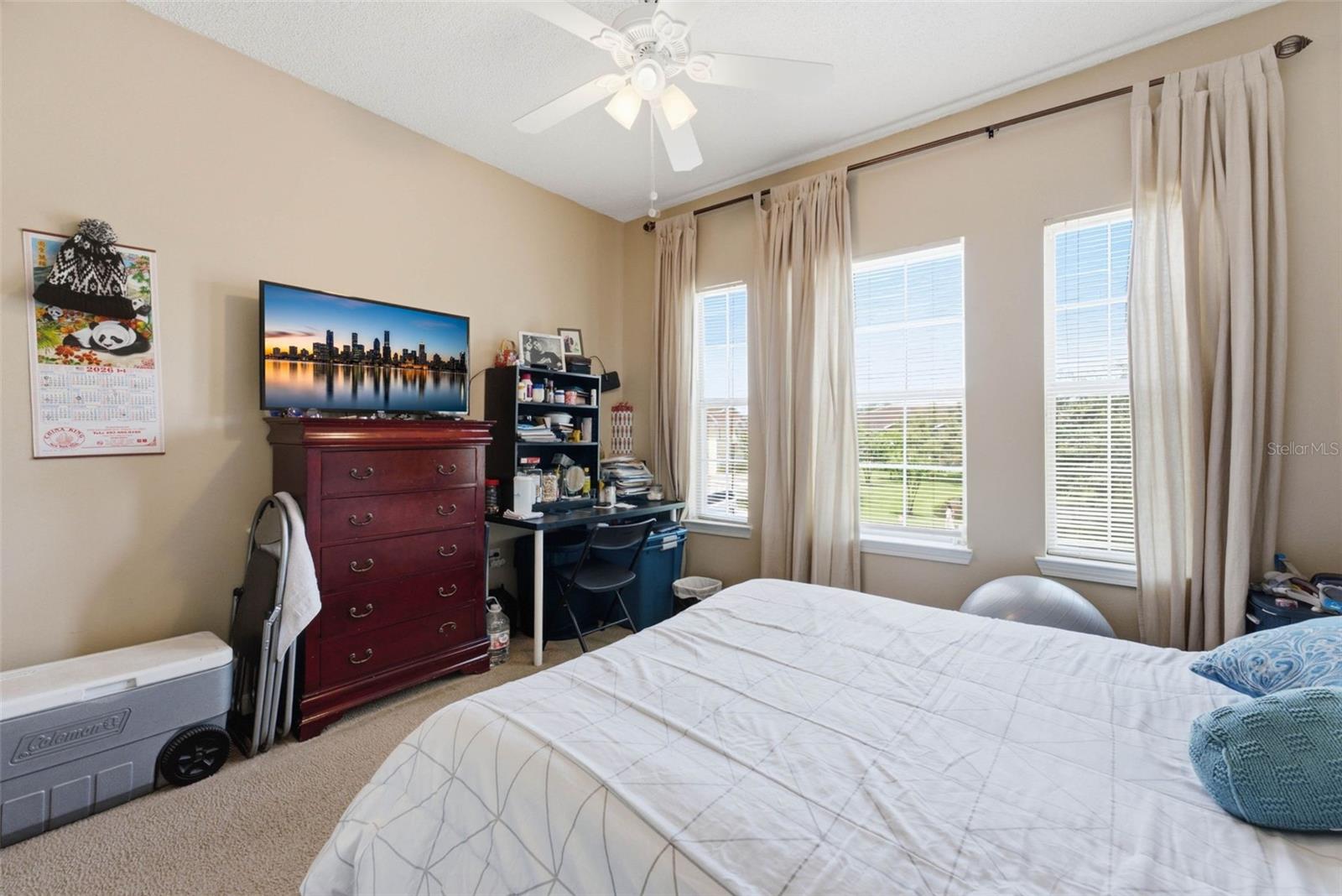 Bedroom with a large bed, three curtained windows, and a desk beside a dark wood dresser.