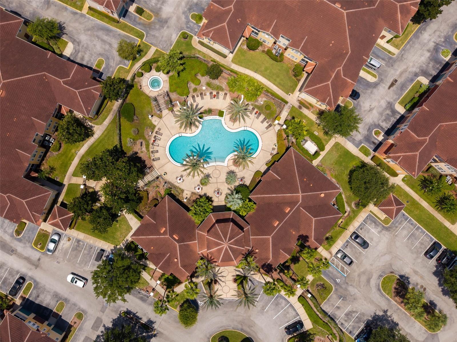 Aerial view of a condominium complex with a curving pool, circular pool, palm trees, and red-tile roofs.
