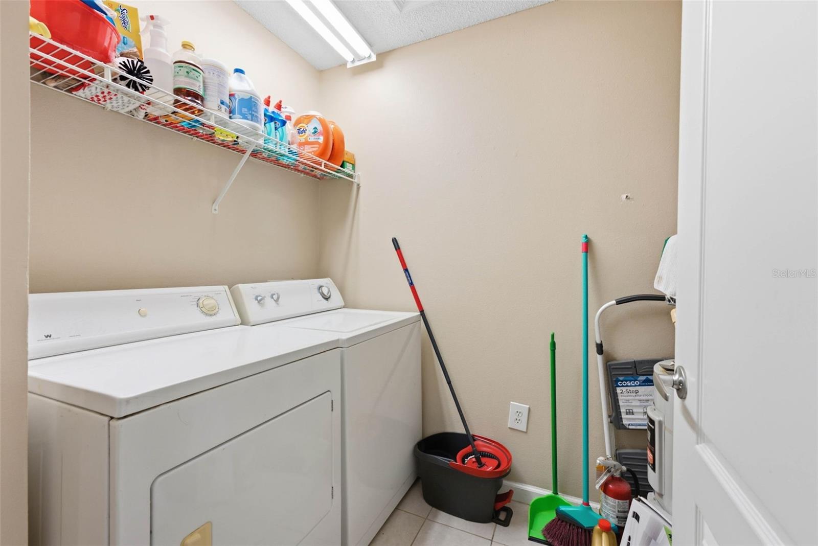 Laundry room with white washer and dryer, cleaning supplies on a wire shelf, and mops by the wall.
