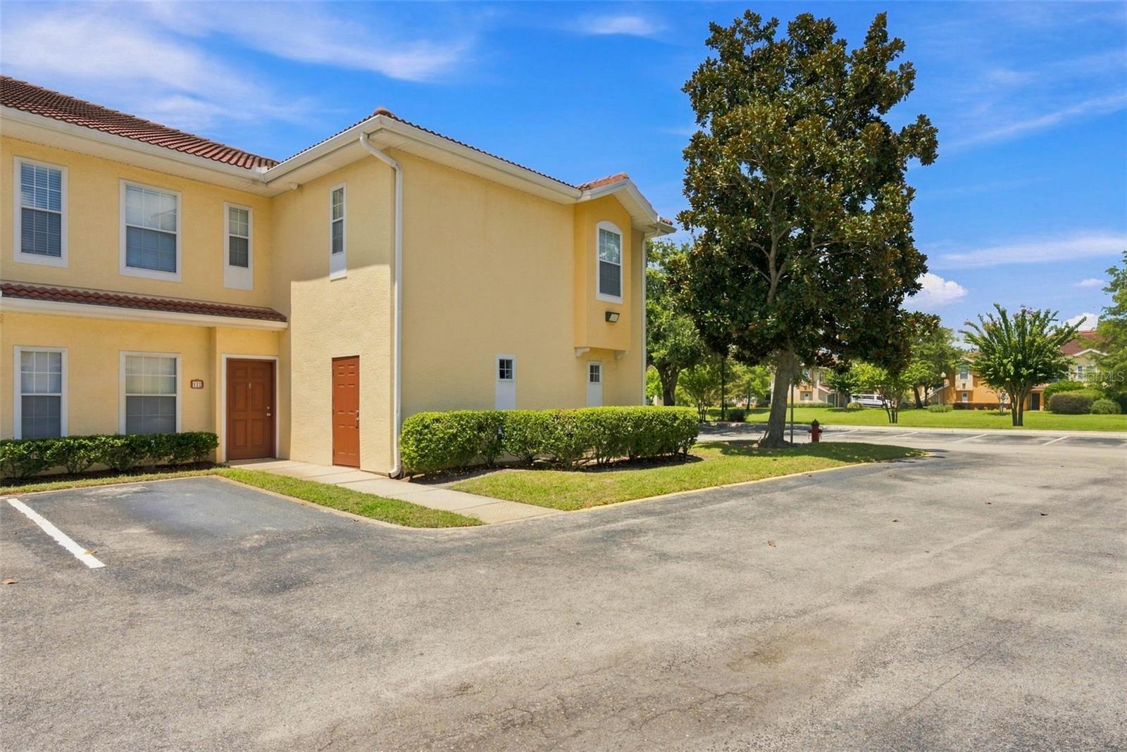 Yellow two-story attached home with terracotta tile roof beside a parking area and large tree.