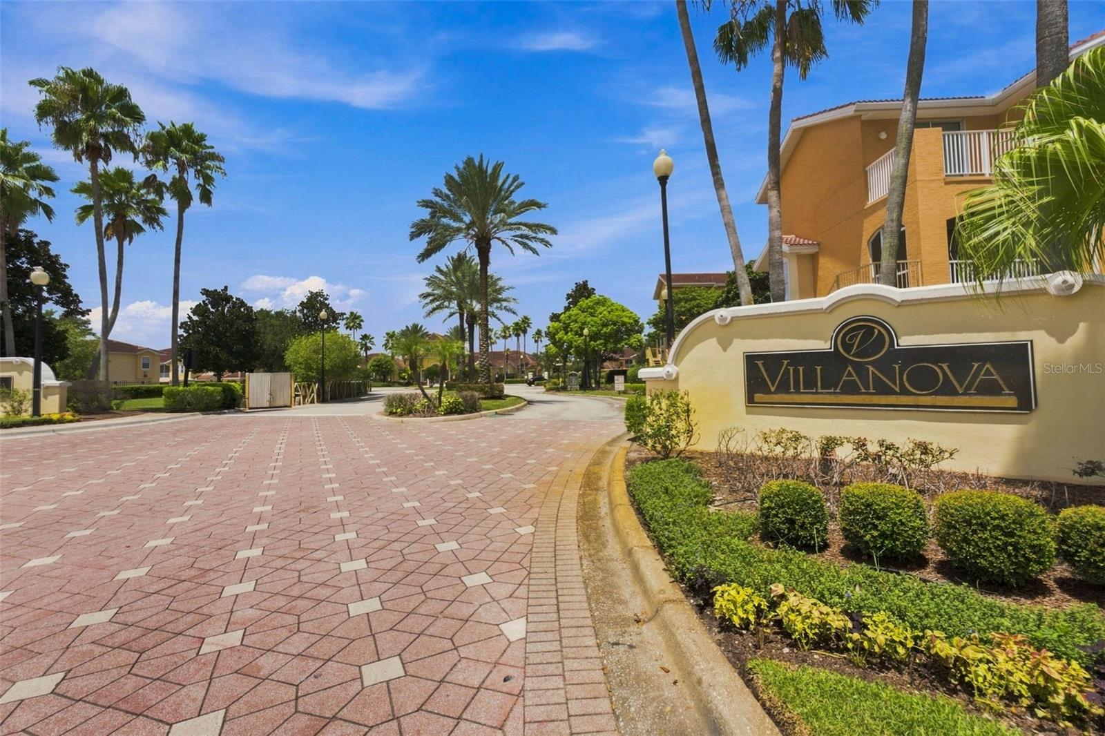 Villanova community entrance with a brick-paved drive, palm trees, and landscaped shrubs.