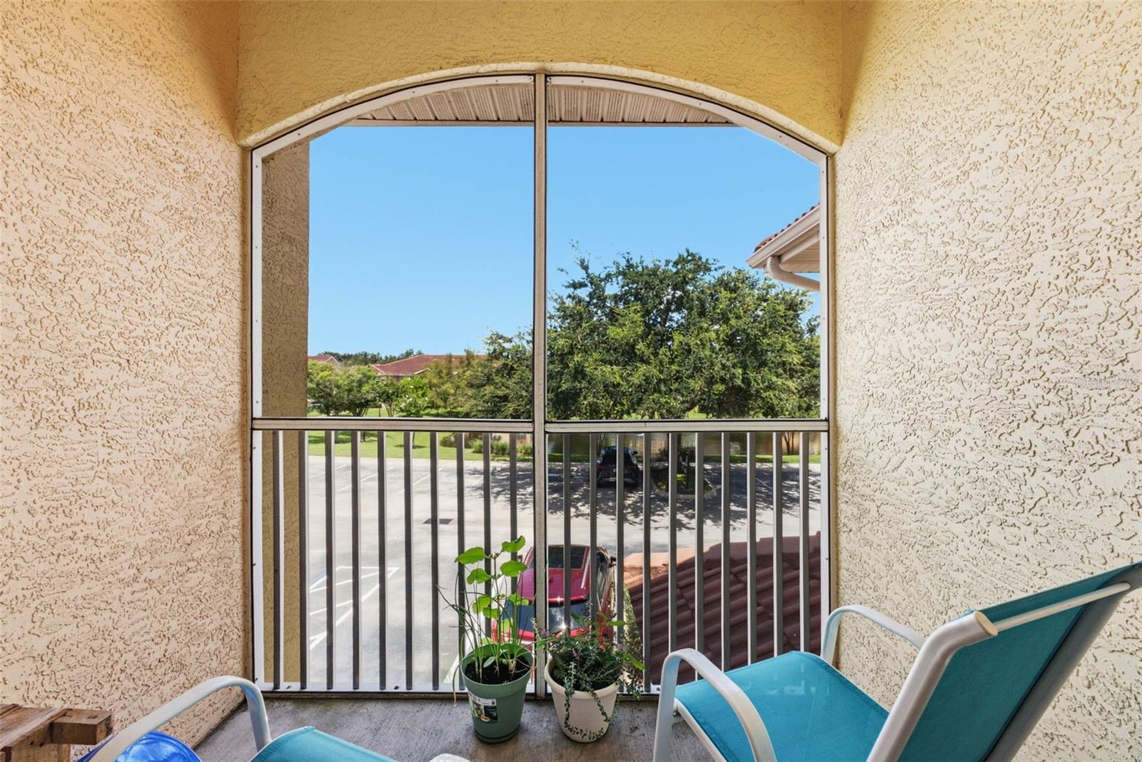 Screened balcony with patio chairs and potted plants overlooking trees and a parking area.