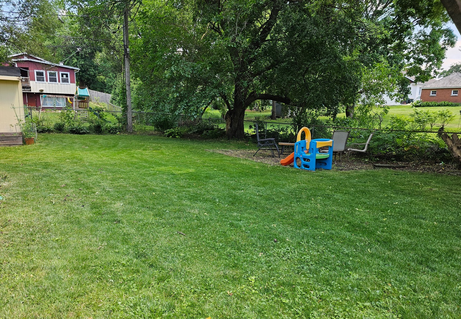 Grassy backyard with a mature shade tree, patio chairs, and a colorful children’s playset.