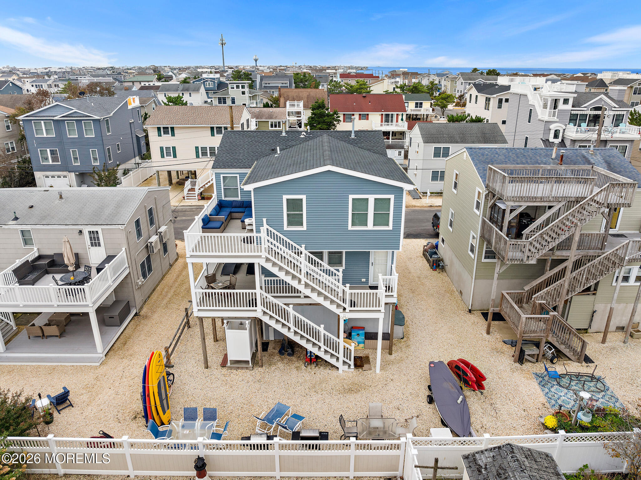 Blue two-story beach house with white decks and exterior stairs, kayaks and paddleboards on the sandy lot.