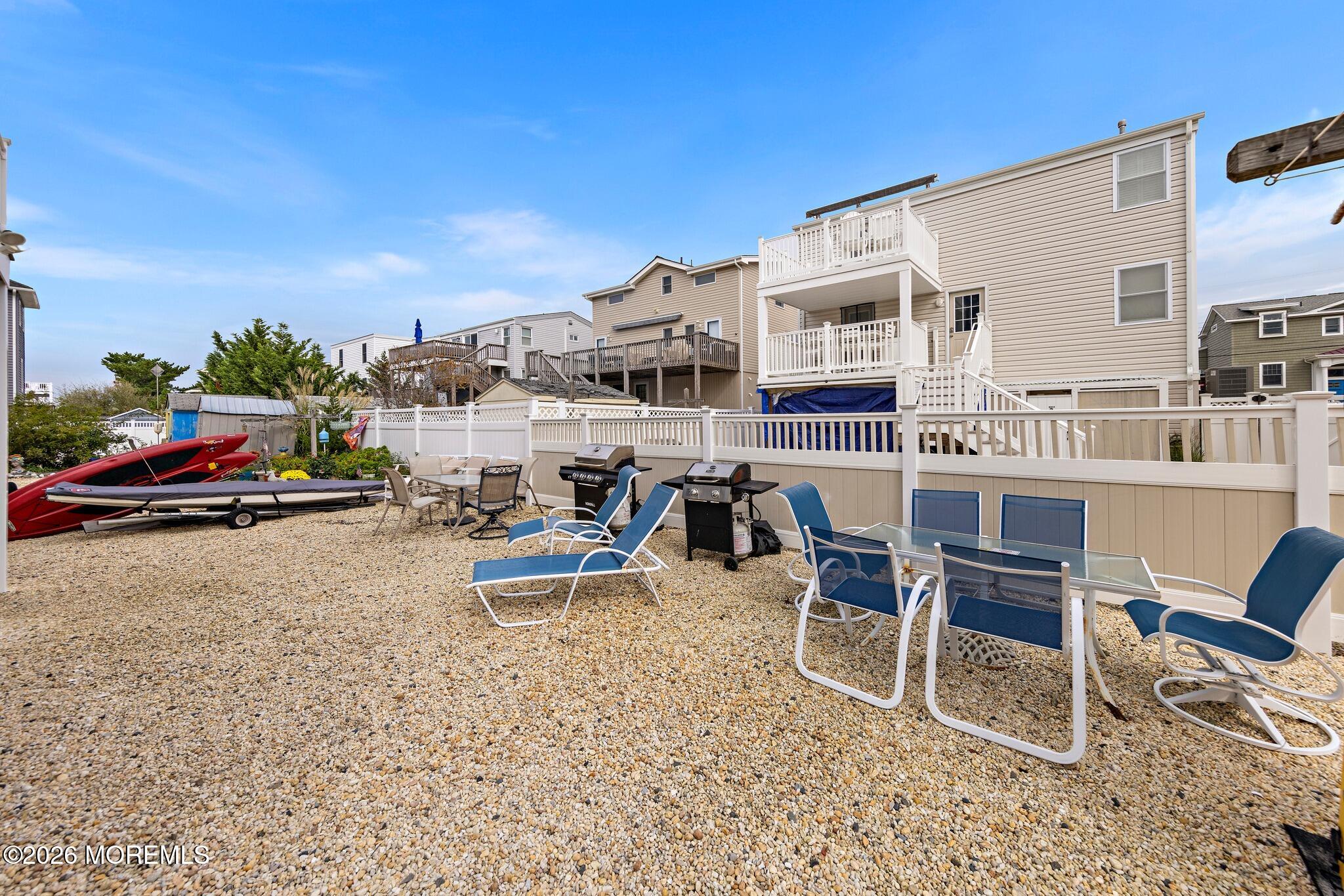 Coastal home rear patio with white balconies, blue lounge chairs, grills, and kayaks on a gravel yard.