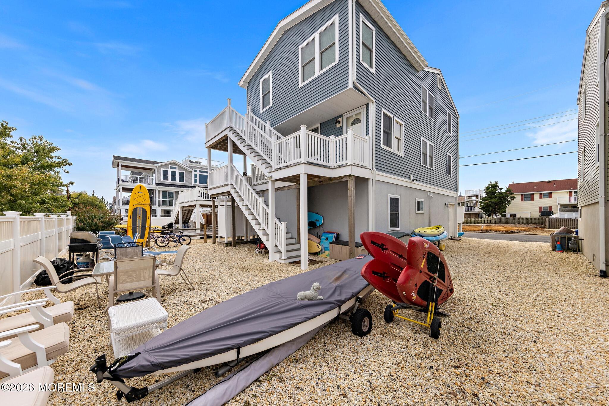 Gray coastal home with white exterior stairs and decks, overlooking a gravel yard with kayaks.