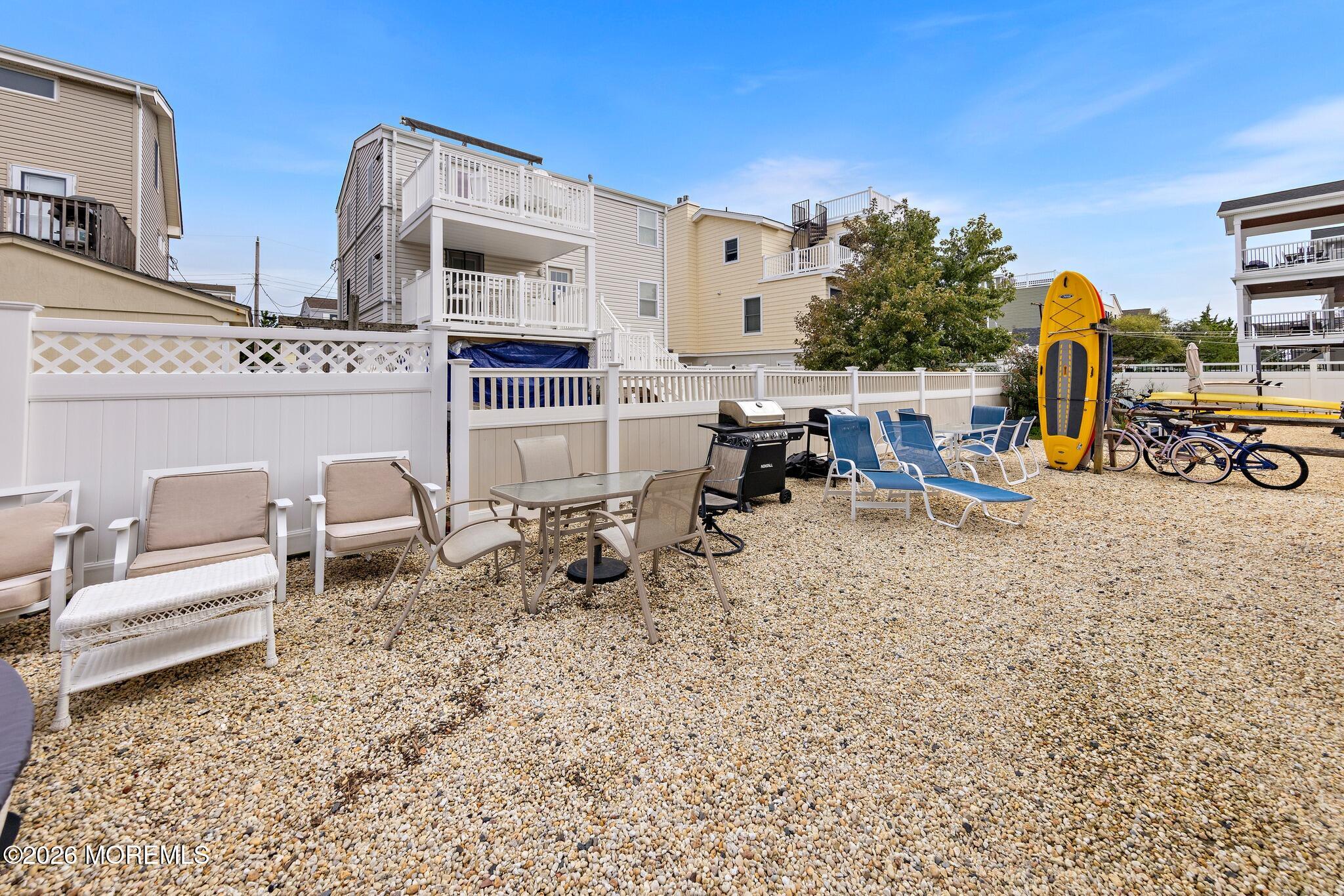 Gravel patio with outdoor seating, a grill, upright kayaks, and bicycles beside coastal homes.