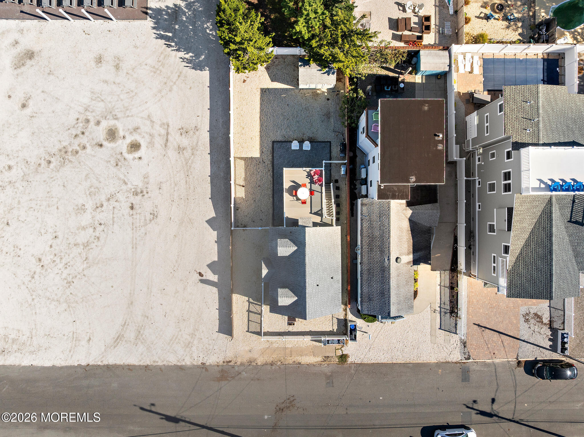 Aerial view of a gray-roofed home with a raised deck beside a large sandy vacant lot.