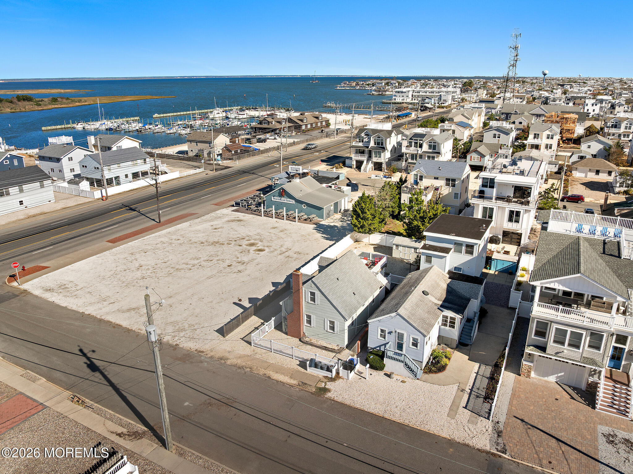 Aerial view of a vacant lot beside a coastal road, with a bay and marina beyond nearby homes.