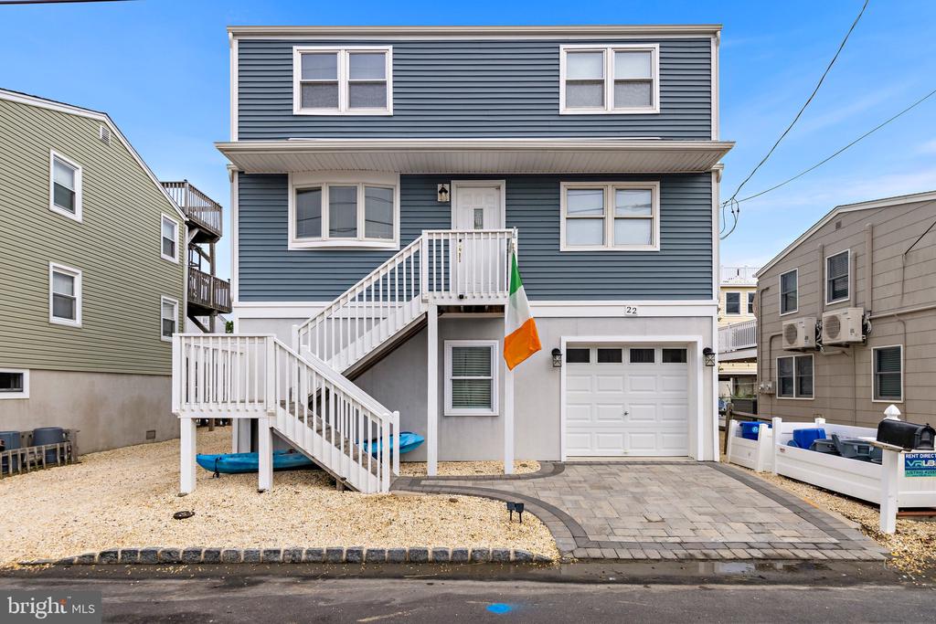 Raised blue coastal home with white exterior stairs, garage, paved driveway, and an Irish flag.