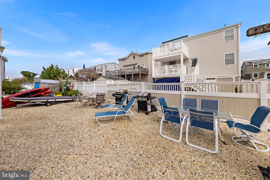 Gravel patio with blue lounge chairs, a dining table, grills, and kayaks beside a beige house.