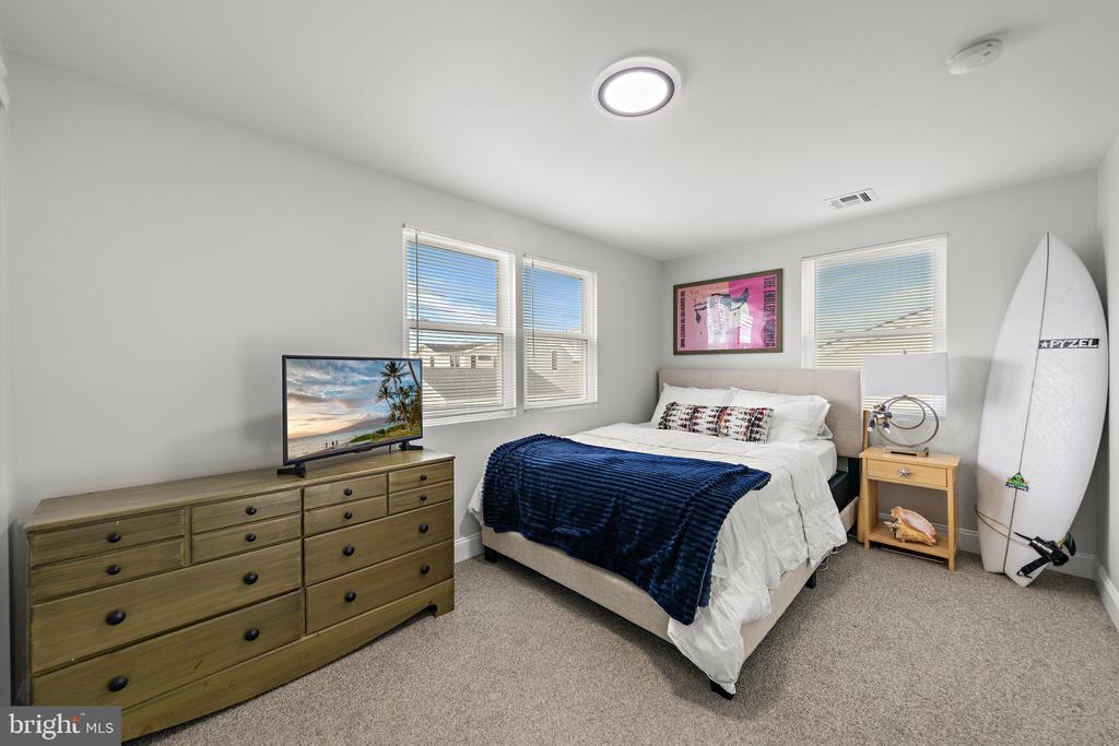 Bedroom with a white bed, navy blanket, wooden dresser, and surfboard by the wall.